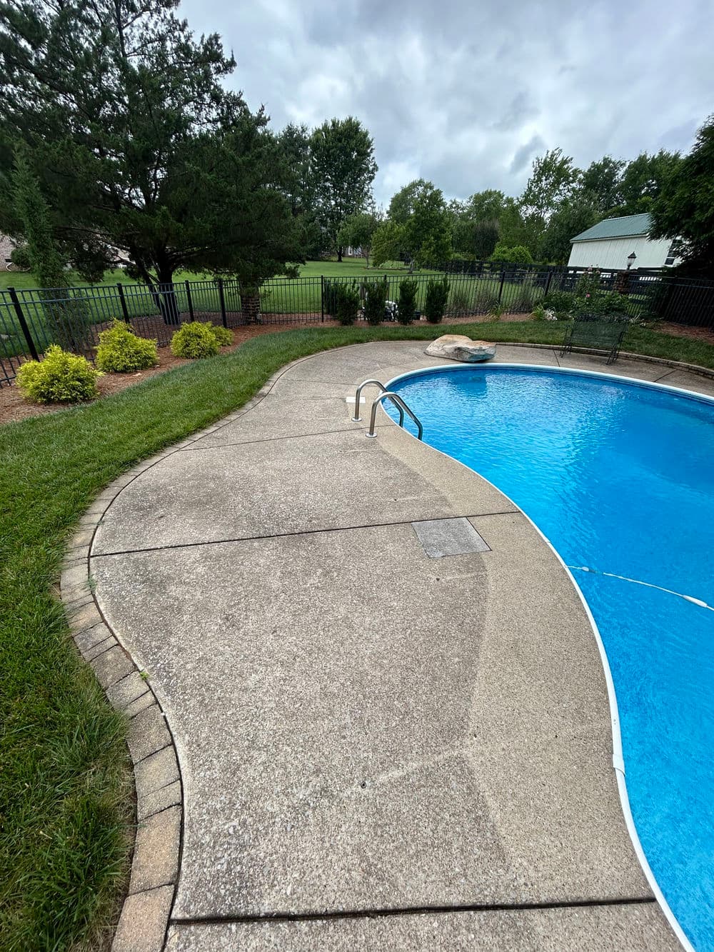 Residential backyard with a swimming pool, concrete deck, and green lawn under cloudy sky.